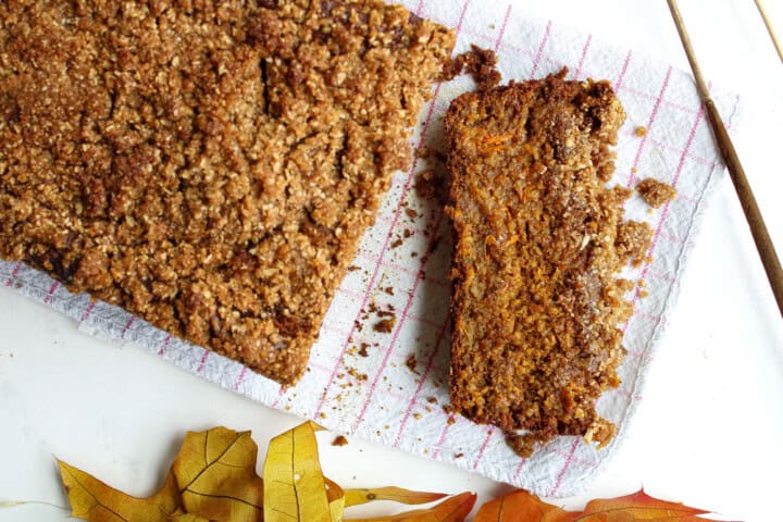 A loaf of pumpkin bread with a streusel topping is partially sliced on a white background with a red-striped cloth. Autumn leaves are scattered nearby for decoration.
